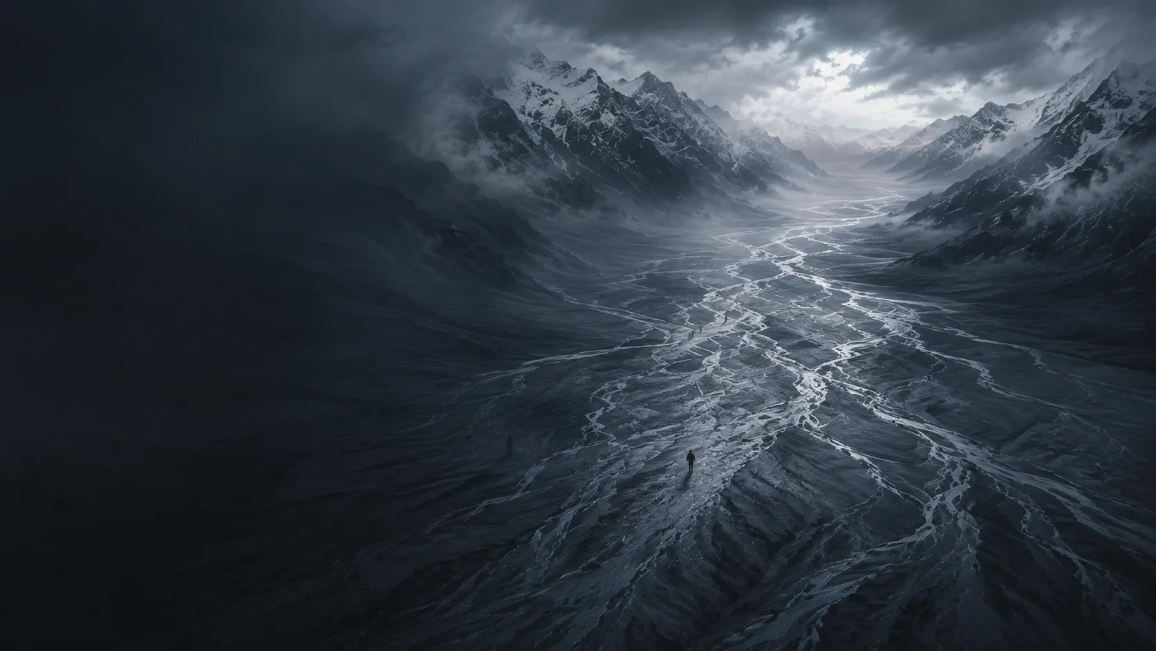 Aerial view of a lone figure standing amid winding braided river channels in a snow-capped mountain valley